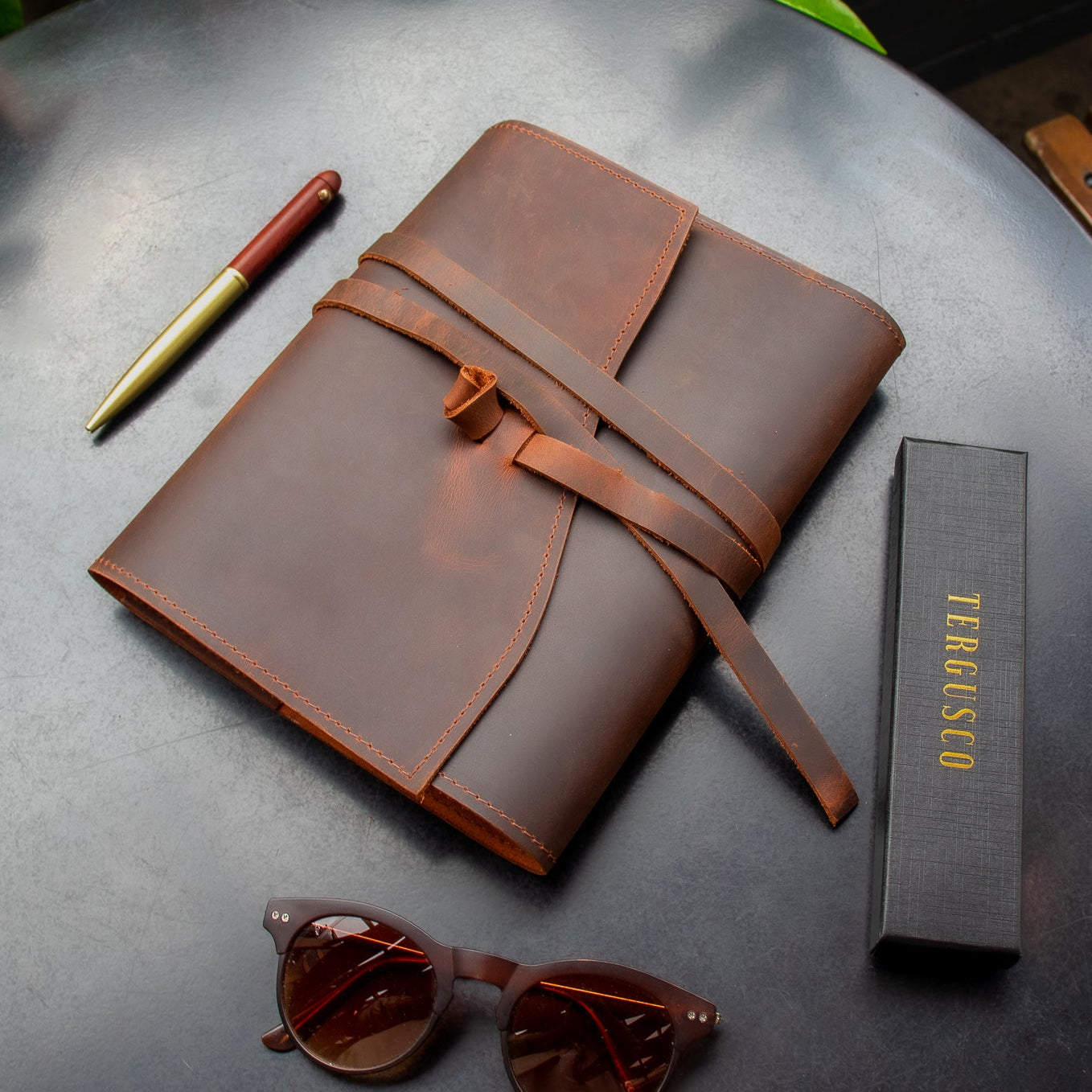 Brown leather notebook with sunglasses and pen on a dark surface with greenery in the background