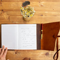Open notebook on a wooden table with a hand, a small plant, and a cork coaster.