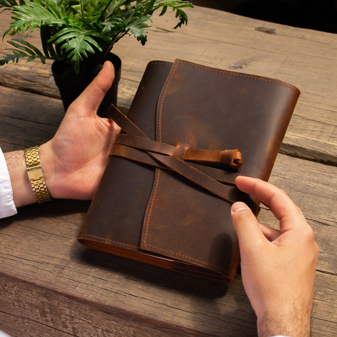 Brown leather notebook held by a person on a wooden surface with a plant in the background