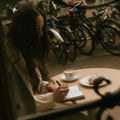 Woman sitting at a table in an outdoor cafe, writing in a notebook with a cup of coffee and pastry.